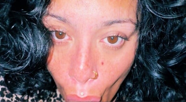 Close-up portrait of a woman with curly dark hair, nose piercing, and expressive face in a dimly lit setting.