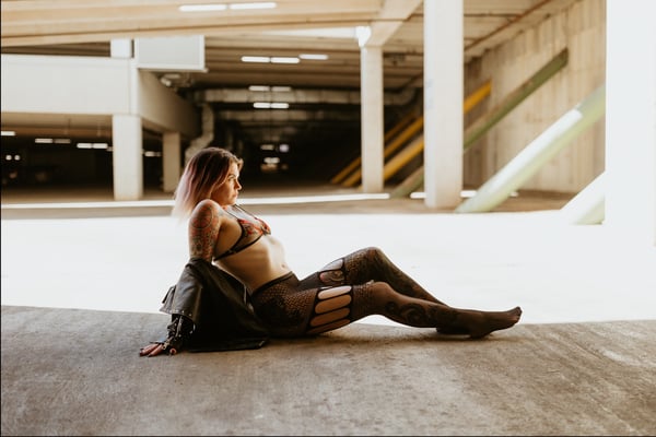 A woman with pink hair and tattoos poses lounging on the floor in a dimly lit parking garage, wearing a jacket, bra, and stockings.