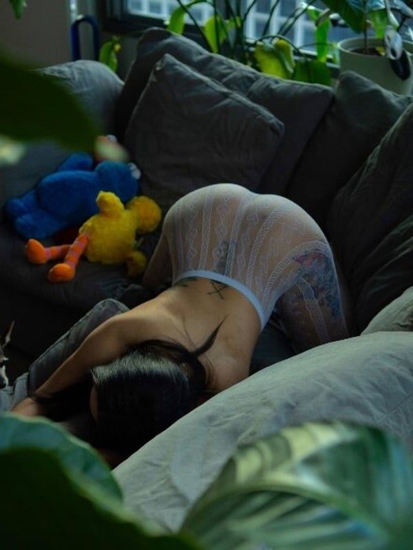 A person lying face down on a couch surrounded by plants, toys, and a cat, wearing a sheer outfit in a cozy living room.