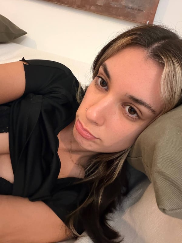 A young woman with highlighted hair lying on a bed, taking a selfie in a low-cut black top, with pillows in the background.