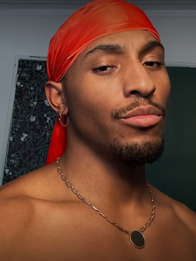 A young man with a red headwrap, earrings, and chain necklace posing shirtless against a patterned background.