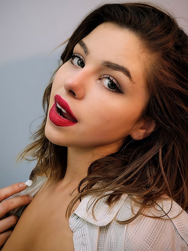 Close-up portrait of a young woman with wavy brown hair, bold red lipstick, and subtle makeup, wearing a white top.