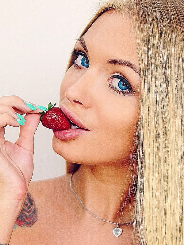 A woman with long blonde hair and blue eyes playfully bites into a strawberry, smiling at the camera.