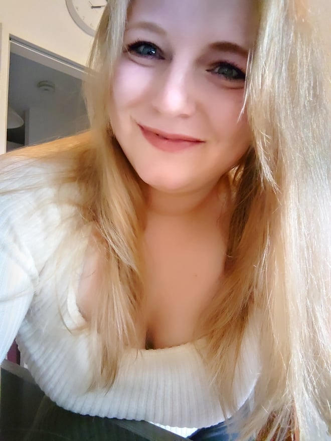 Young woman with long blonde hair smiling at the camera indoors, wearing a white sweater.