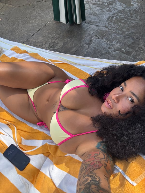 A woman in a yellow and pink bikini taking a selfie while lying on a striped towel outdoors.
