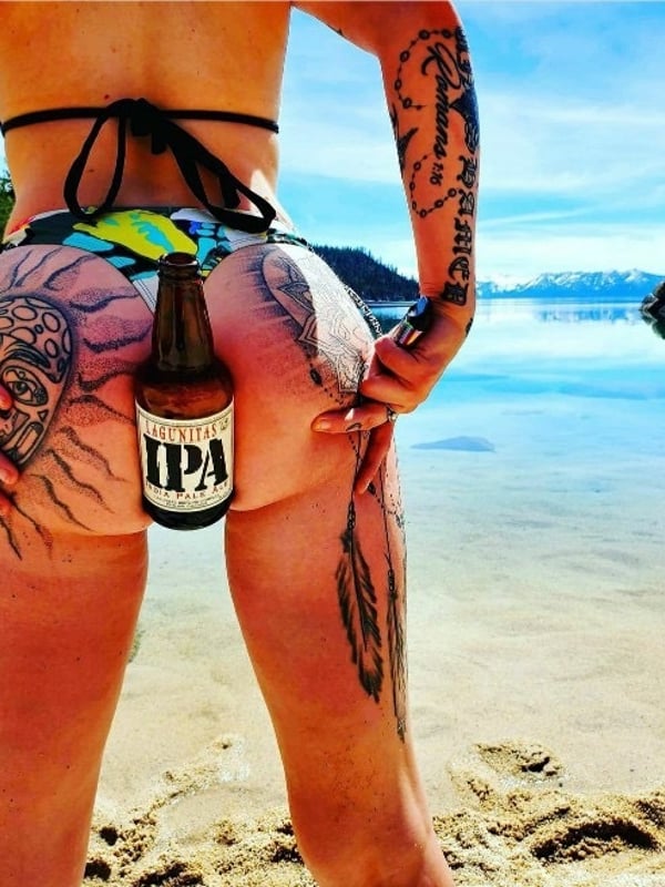 A woman in a colorful bikini standing by a lake, holding a beer bottle near her hip with tattoos visible on her arms and lower back.