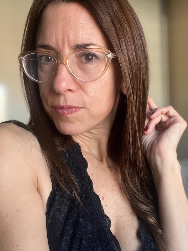 A woman with glasses and long brown hair, wearing a black lace top, gazing directly at the camera indoors.
