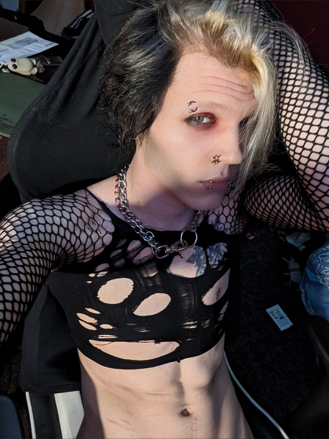 Person with split-colored hair and piercings wearing fishnet sleeves and chains, sitting in a casual setting.