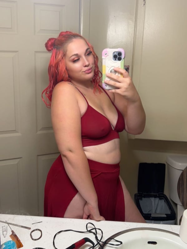 Young woman with red hair in pigtails taking a mirror selfie in a bathroom, wearing a red bra and skirt set.