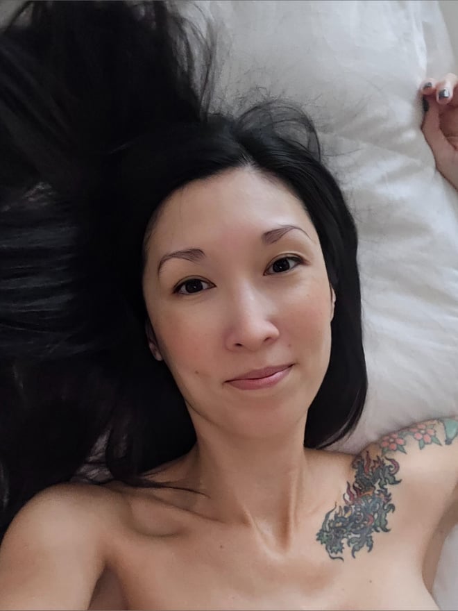 A woman with long dark hair and tattoos lying on a bed, smiling at the camera with pillows in the background.