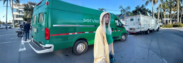 A young woman with long blonde hair wearing a yellow hooded cape stands on a sunny street next to green vans and palm trees.