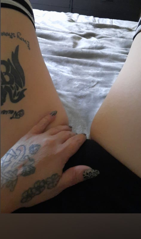 Close-up view of a person's legs and hands with intricate tattoos, posed on a bed with dark shorts.