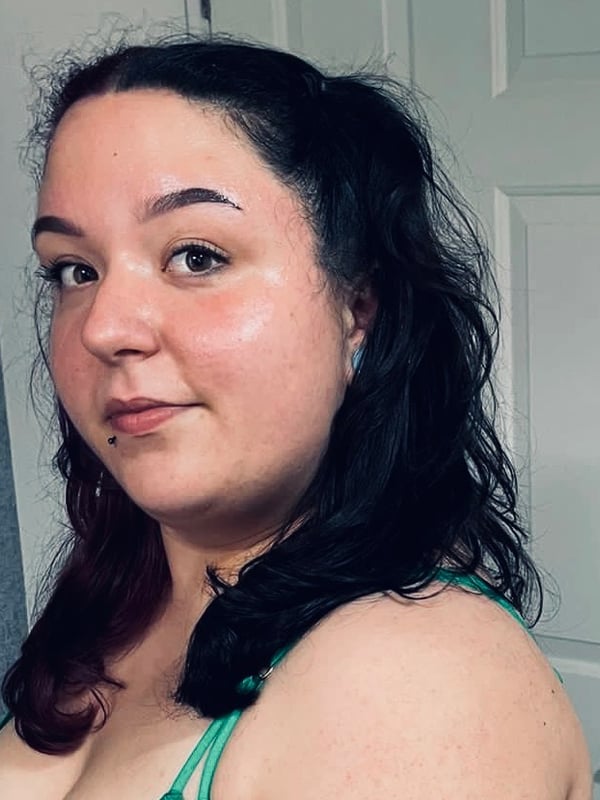 A woman with dark wavy hair in pigtails smiling at the camera in a green top inside a room.