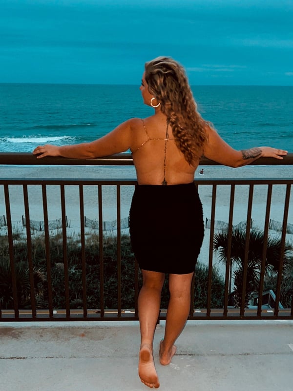 Woman standing on a balcony overlooking the ocean at dusk, wearing a backless top and skirt with arms extended on the railing.