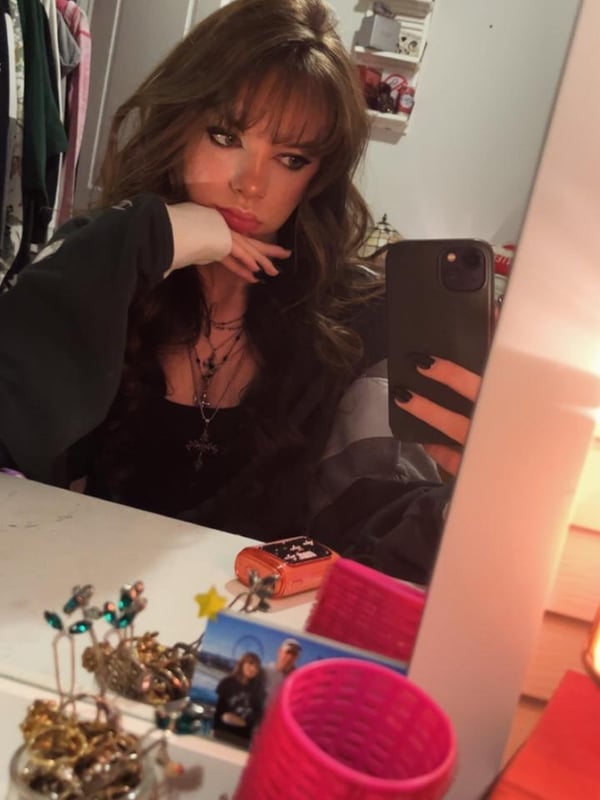 A person taking a mirror selfie wearing a black dress, with long brown hair and visible tattoos on the arm.