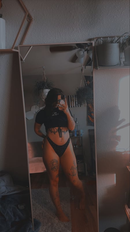 Young woman standing in front of a mirror in a cozy room with plants and geometric decor, wearing a cropped top and shorts.