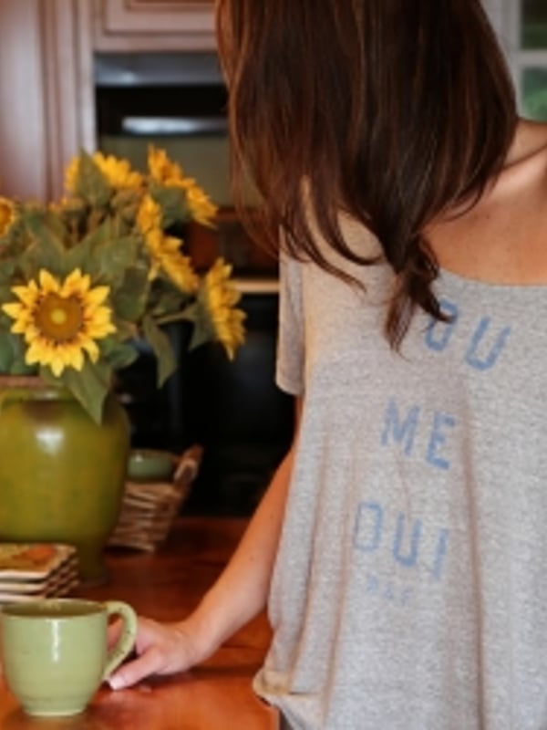 Woman with long brown hair standing in a kitchen, holding a mug, wearing a loose gray top next to sunflowers and wooden decor.