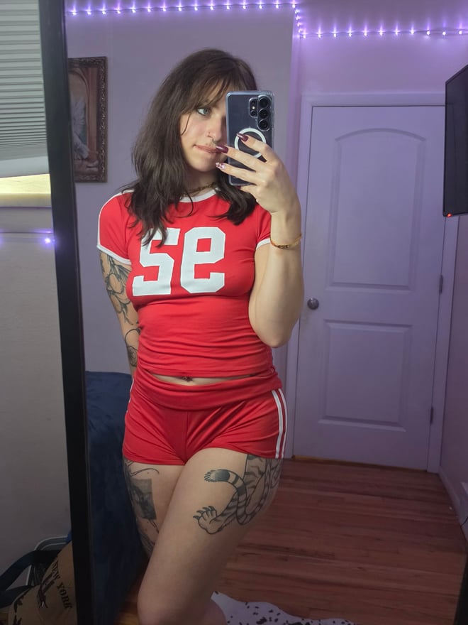A brunette woman with facial piercings and tattoos takes a playful mirror selfie while wearing red clothing.