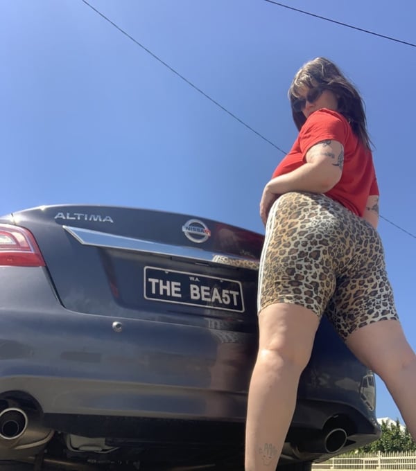 A woman in a red shirt and leopard print shorts posing next to a gray Nissan Altima car labeled 'THE BEAST' under a clear blue sky.