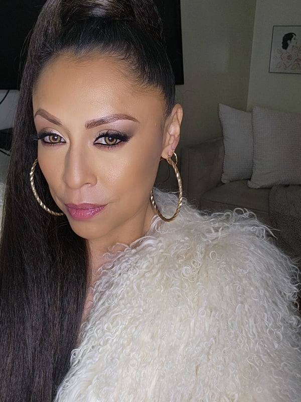 A woman with long dark hair in a ponytail, wearing large hoop earrings and a fluffy white jacket, posing indoors with a confident expression.