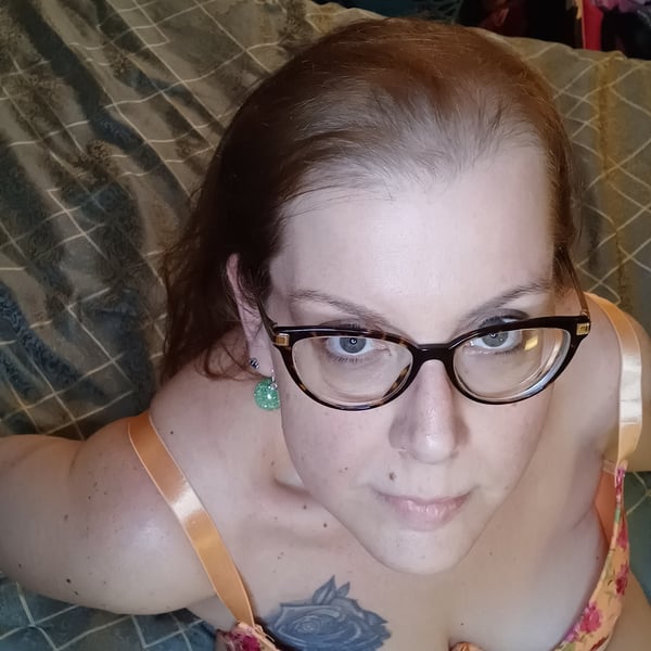 A woman with glasses and brunette hair posing on a bed, wearing an orange top, with tattoos visible on her chest.