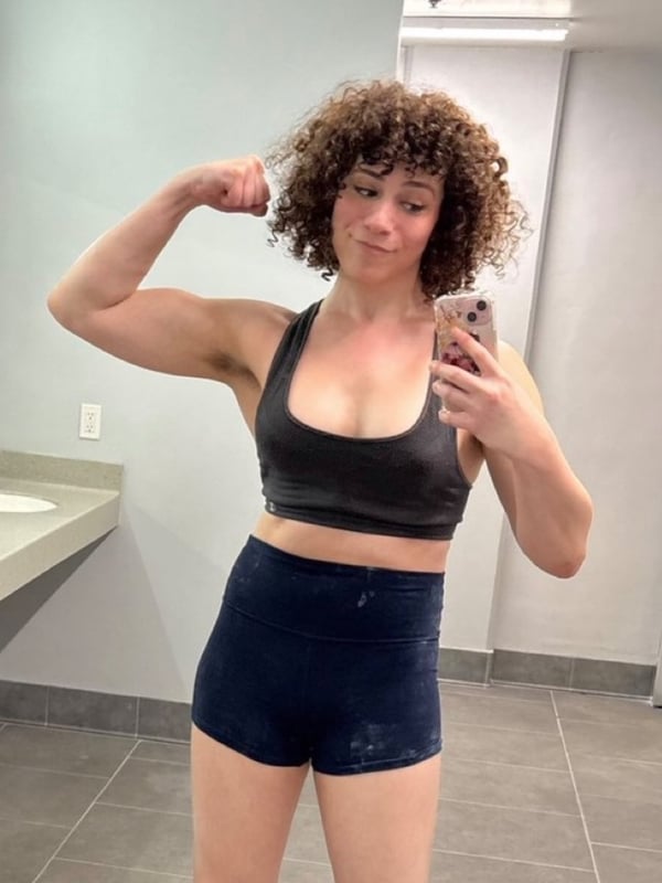 A person with curly hair flexing arm muscles in a black tank top and shorts, smiling in a bathroom mirror selfie.