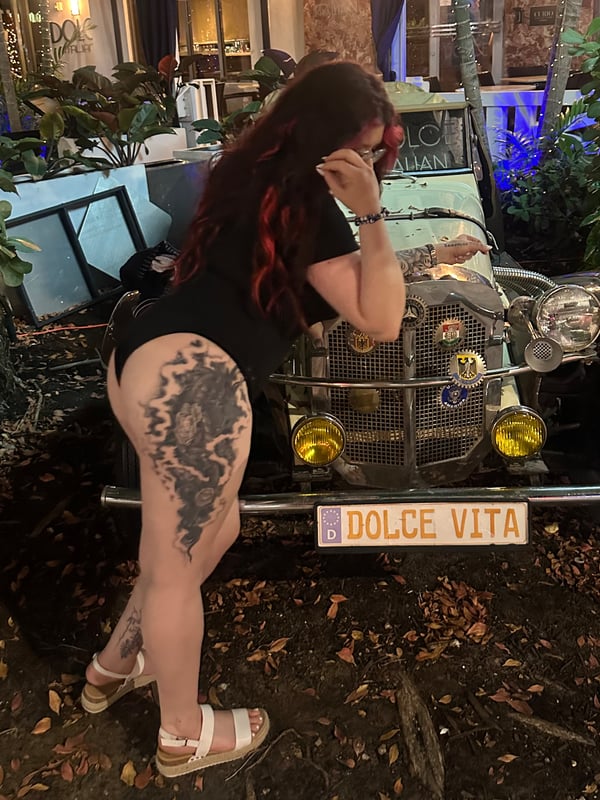 Young woman with red hair leaning against a vintage car in an outdoor setting with plants and warm lighting.