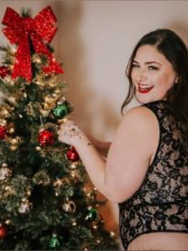 Woman smiling next to a decorated Christmas tree, dressed in a lace top with holiday ornaments nearby.