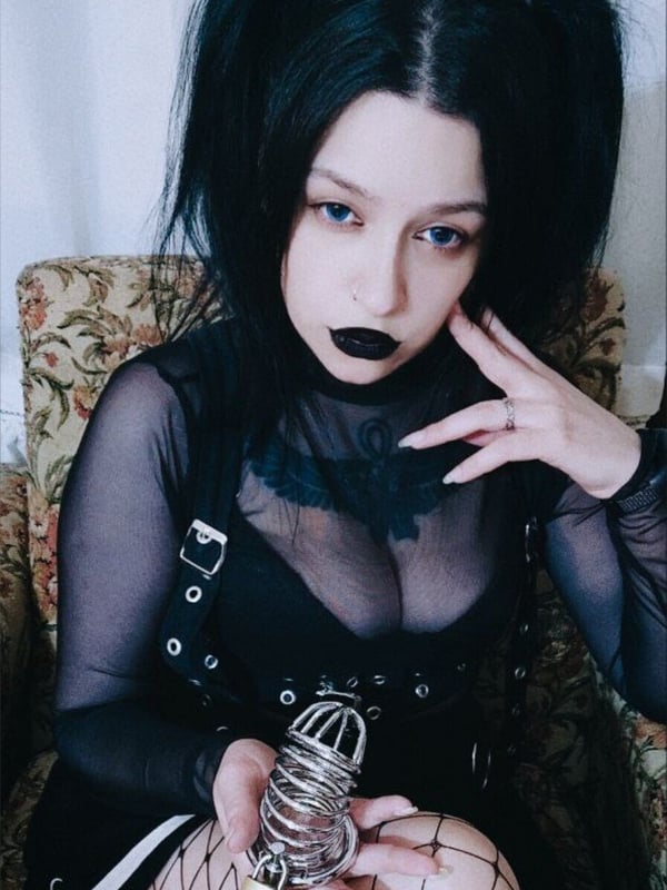A woman with black hair in pigtails and bold black lipstick sits holding a metal cage prop, dressed in sheer black gothic clothing on a patterned chair.