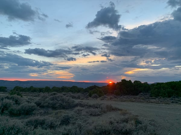 Scenic sunset over a rugged outdoor landscape with bushes, mountains, and a dirt path under a cloudy sky.