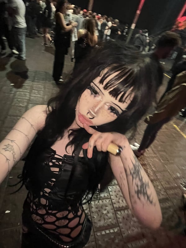 A young woman with bold makeup and piercings stands outdoors near a train station, dressed in a dark furry coat with long black hair.
