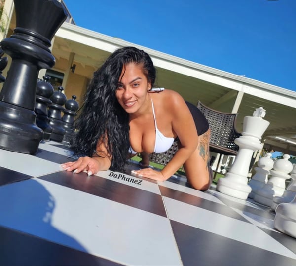 Woman with long dark wavy hair smiling and posing playfully on a giant chessboard outdoors next to large chess pieces, wearing a white bikini top and black bottoms.