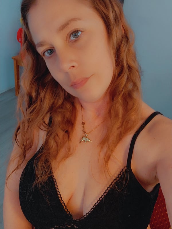 Woman with wavy reddish-brown hair taking a selfie in a lace top, standing indoors with a neutral expression.