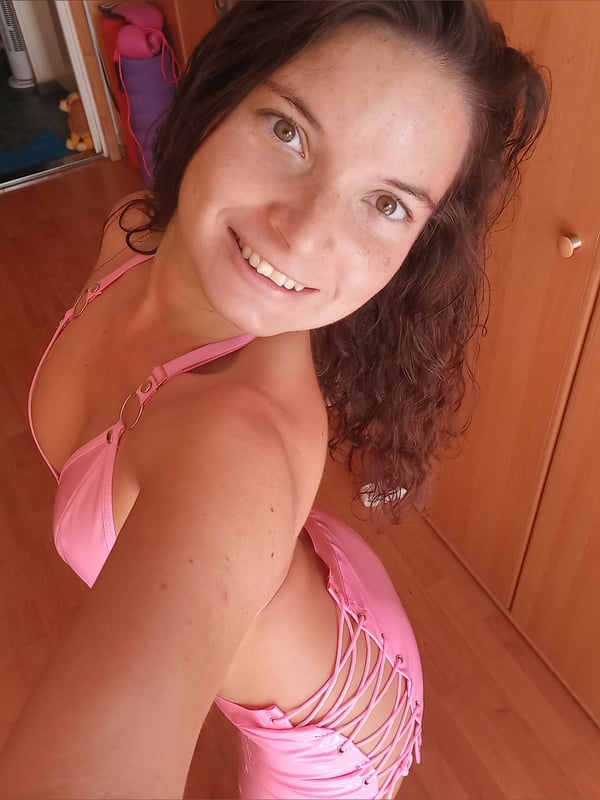 A woman with curly brown hair smiling, wearing a pink strap bikini, standing in a room with wooden floors and pink exercise equipment.