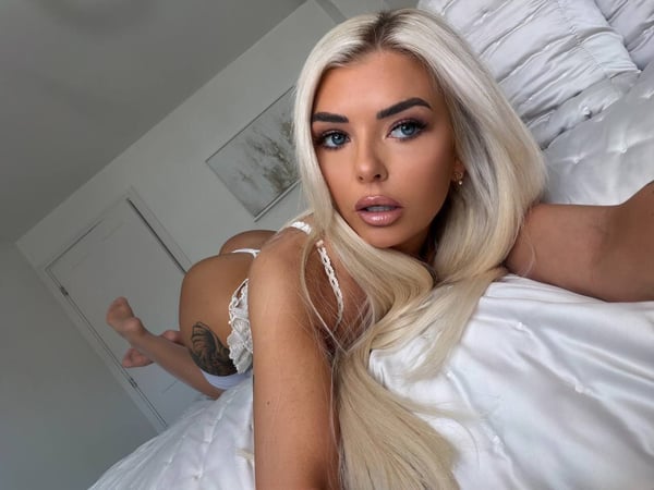 A woman with long blonde hair posing on a white bed in a bright bedroom.