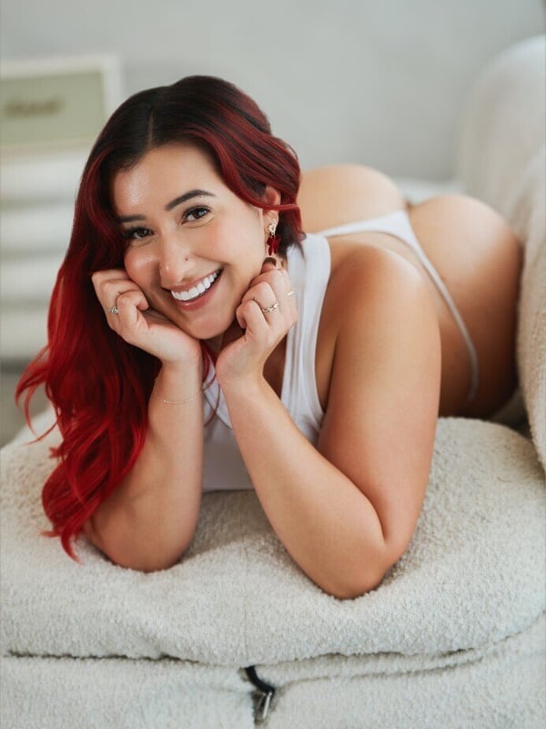 A smiling woman with red hair lying on a white couch in light-colored lingerie, posing playfully indoors.