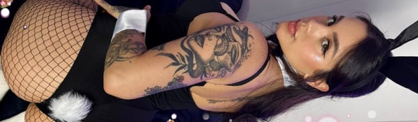 A woman with long dark hair and tattoos posing in a black outfit with fishnet stockings and bunny ears.
