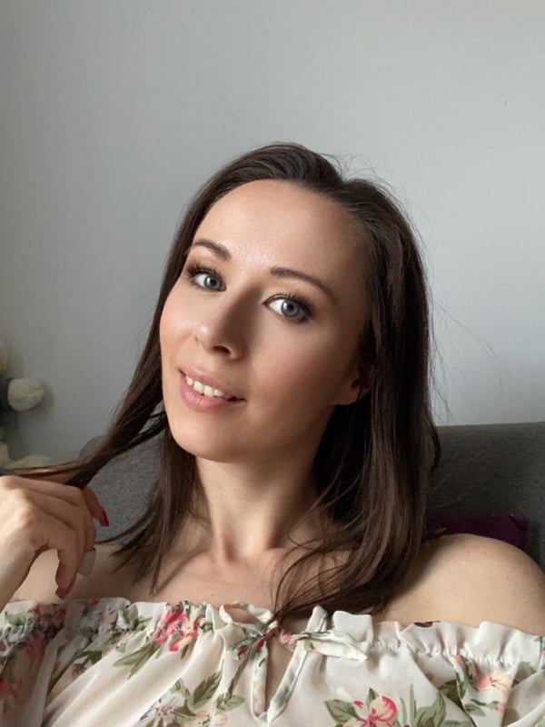 A young woman with long brown hair smiling at the camera, wearing a floral off-shoulder top while seated indoors.