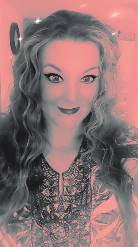 A woman with long wavy blonde hair smiling at the camera, wearing a patterned top, with a pink-toned background and decorative elements.