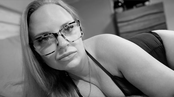 A woman with blonde hair and glasses lying on a bed, wearing a black top, in a black and white photo.