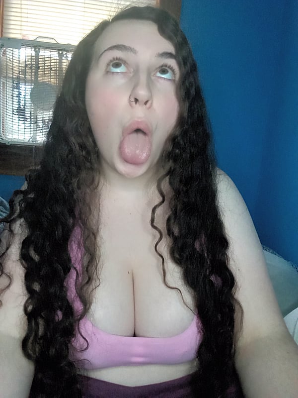 A woman with long curly dark hair sticking out her tongue, wearing a pink top, sitting in a room with blue walls and window blinds.