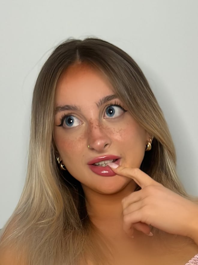 Young woman with blonde hair and freckles posing with hand near mouth, wearing hoop earrings against a plain background.