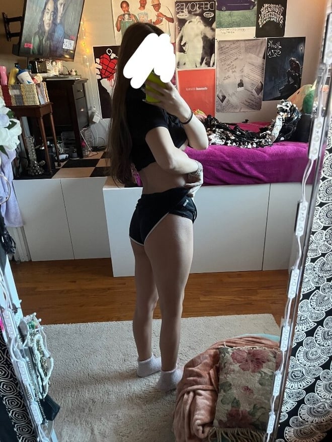 Young woman with long hair standing in front of a mirror in a colorful bedroom with posters and furniture.