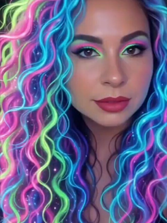 A woman with vibrant neon-colored curly hair in shades of green, blue, and pink, posing with bold makeup and a confident expression.