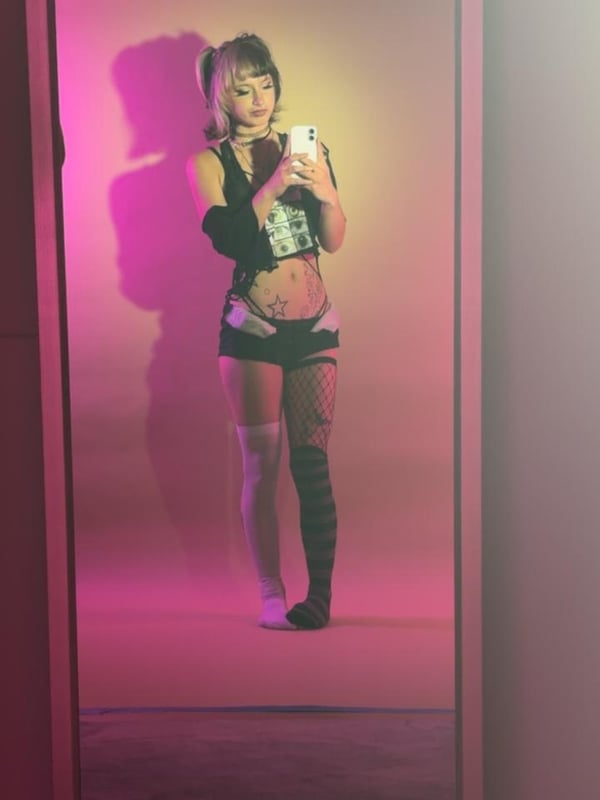 A young woman sitting on stairs in fishnet stockings, black boots, and a black top with tattoos visible, posing confidently outdoors.