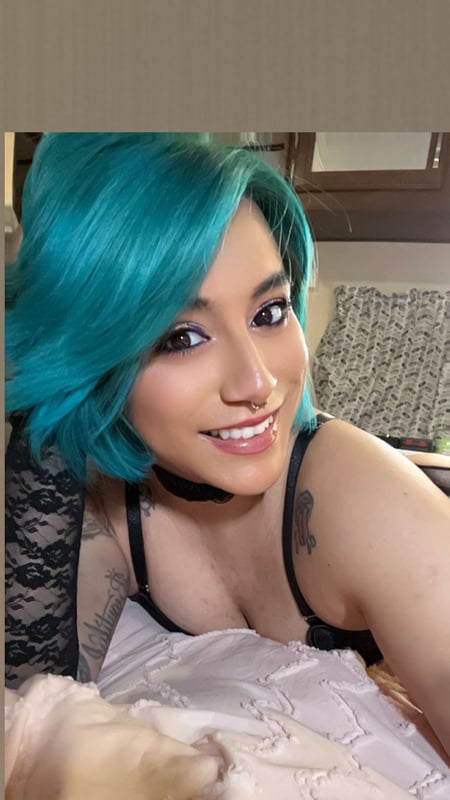 A woman with turquoise dyed hair wearing lace lingerie and a choker, smiling in an indoor setting with curtains in the background.