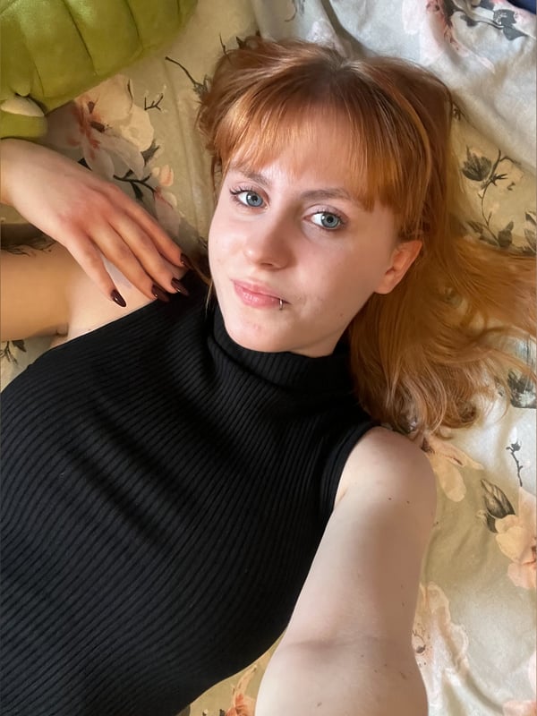 A young woman with red hair and blue eyes smiling at the camera, lying on a bed wearing a black top.