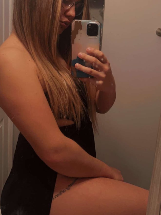 A young woman taking a mirror selfie in a bathroom, wearing a sheer black top and sitting on the floor.