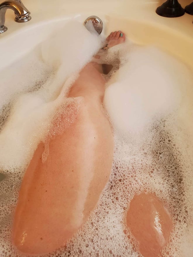 Close-up view of legs and feet relaxing in a foamy bathtub with chrome faucet in the background.