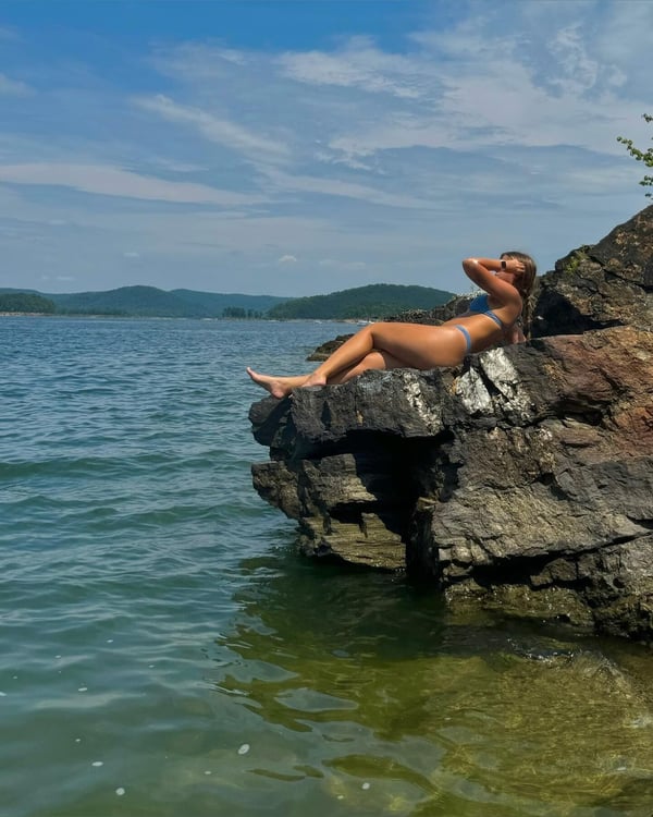 A woman lounging on a rock by the water in a bikini, with mountains and a lake in the background under a blue sky.